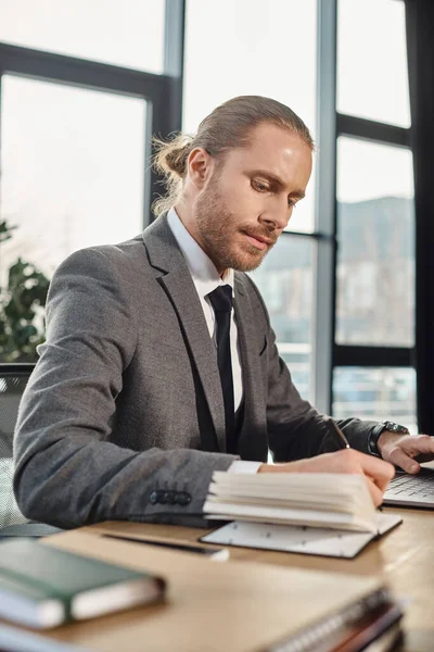 Homme d'affaires concentré en costume gris écrit dans un ordinateur portable près d'un ordinateur portable dans un environnement de travail moderne — Photo de stock