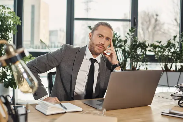 Sério homem de negócios elegante sentado no local de trabalho perto do laptop e olhando para a câmera no escritório — Fotografia de Stock