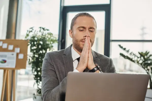 Worried businessman sitting with praying hands and looking at laptop while working in office — Stock Photo