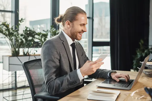 Alegre hombre de negocios en traje gris que sostiene el teléfono inteligente mientras se utiliza el ordenador portátil en el lugar de trabajo en la oficina - foto de stock