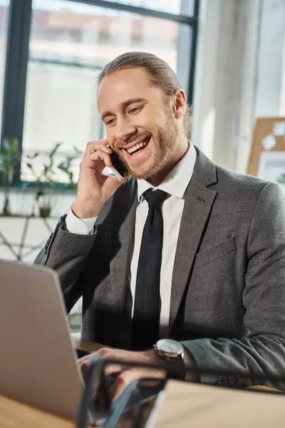 Joyful stylish businessman talking on smartphone near laptop at workplace in modern office — Stock Photo