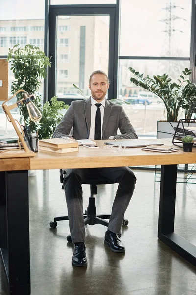 Homme d'affaires confiant assis au bureau avec des documents et des appareils numériques dans le bureau moderne — Photo de stock