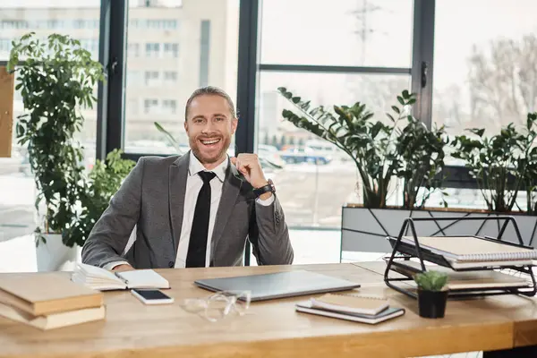 Cheerful businessman looking at camera at work desk with documents and digital devices in office — Stock Photo