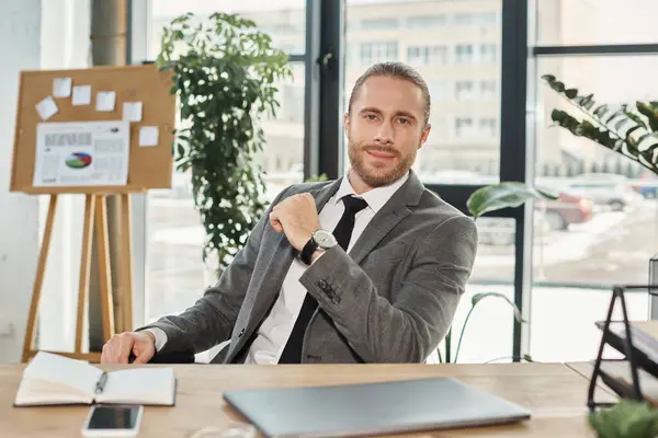 Uomo d'affari ambizioso positivo in abito grigio guardando la fotocamera vicino al laptop e allo smartphone in ufficio — Foto stock