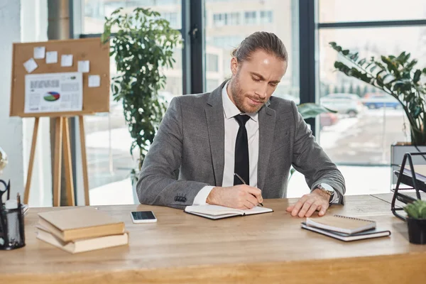 Ocupado hombre de negocios en la escritura de ropa formal en el portátil cerca de teléfono inteligente en el escritorio en la oficina moderna - foto de stock