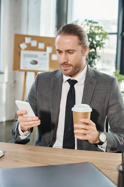 Élégant homme d'affaires en costume gris tenant boisson à emporter et messagerie sur smartphone au bureau — Photo de stock