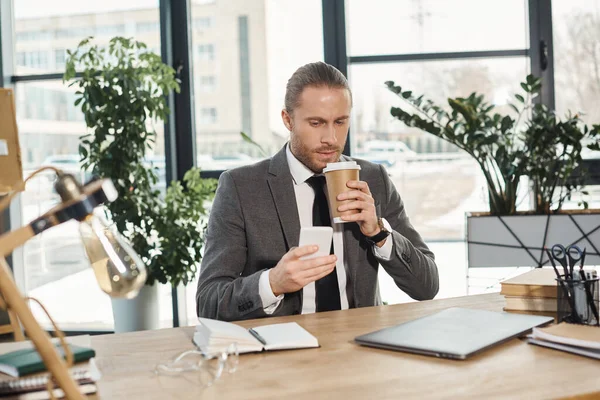 Homme d'affaires branché en costume gris boire du café pour aller et discuter sur smartphone au bureau — Photo de stock