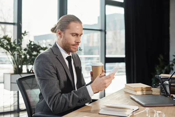 Serious businessman with smartphone and coffee to go near laptop and notebooks on work desk — Stock Photo
