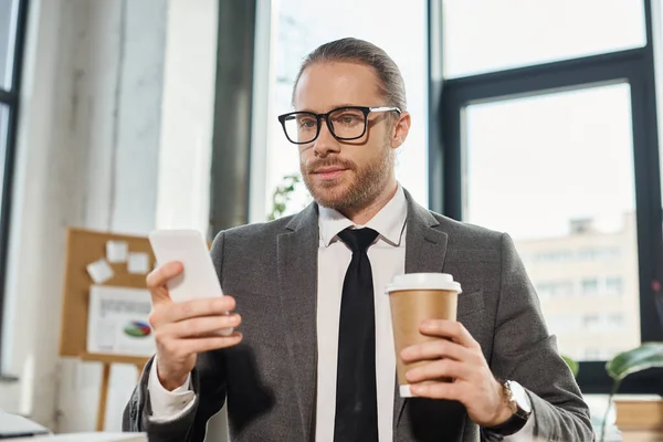 Uomo d'affari in abito grigio e occhiali con cellulare e drink da asporto seduto in ufficio — Foto stock