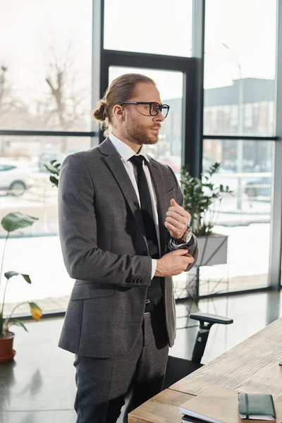 Successful businessman in eyeglasses and grey suit standing and looking away in contemporary office — Stock Photo