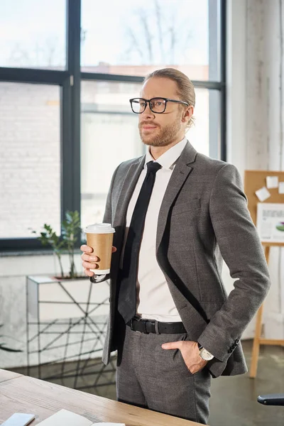 Homem de negócios elegante em óculos segurando café para ir enquanto de pé com a mão no bolso no escritório — Fotografia de Stock