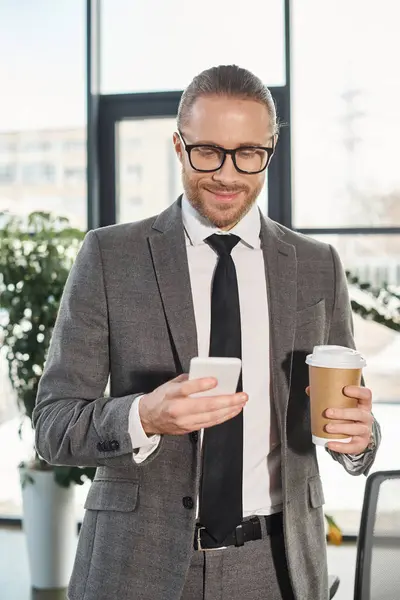 Homme d'affaires souriant en costume gris et lunettes avec café à emporter et téléphone portable au bureau — Photo de stock
