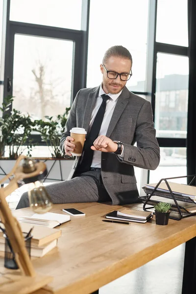 Uomo d'affari con caffè per andare a guardare l'orologio da polso seduto sulla scrivania in un ufficio moderno — Foto stock