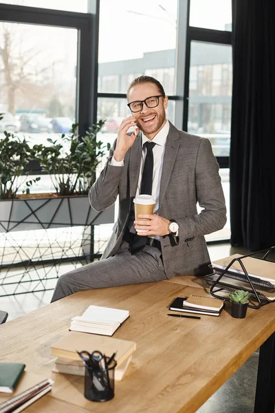 Joyful businessman in formal wear talking on smartphone while sitting on work desk in office — Stock Photo