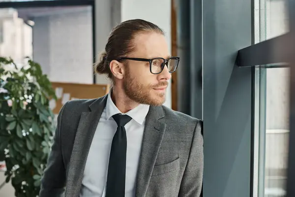 Portrait of thoughtful businessman in grey suit and eyeglasses standing and looking away in office — Stock Photo