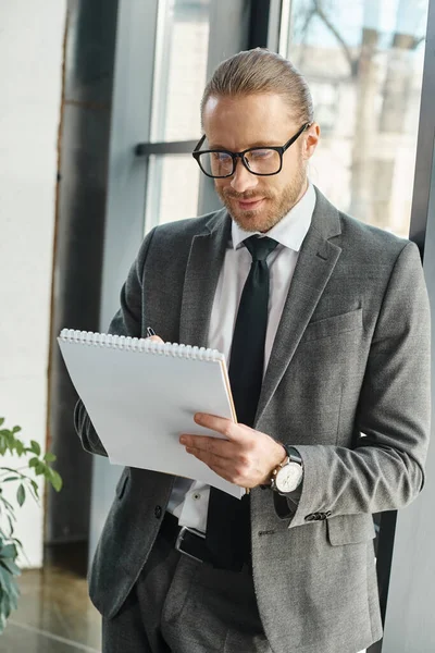 Uomo d'affari sorridente in abito grigio e occhiali da vista che scrive sul taccuino mentre è in carica — Foto stock