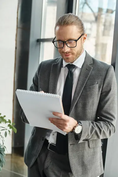 Uomo d'affari sorridente in abito grigio e occhiali da vista che scrive sul taccuino mentre è in carica — Foto stock