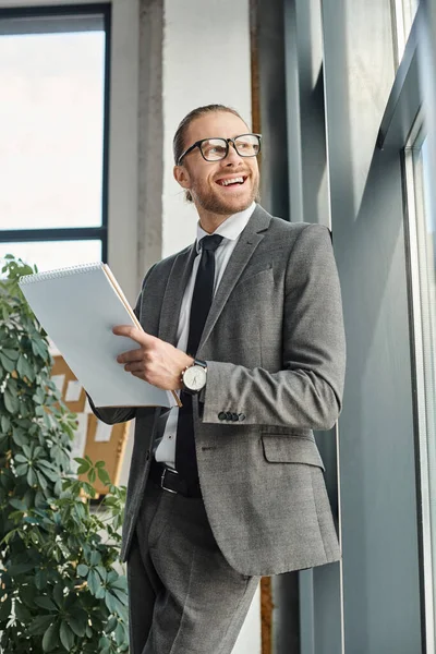 Joyful businessman in grey suit and eyeglasses holding notebook and looking away in office — Stock Photo
