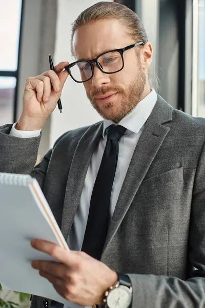 Thoughtful businessman in grey suit adjusting eyeglasses and looking at notebook in modern office — Stock Photo
