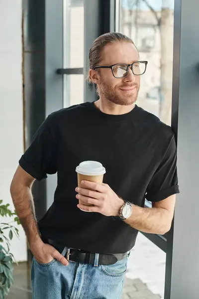 Smiling manager in black t-shirt and eyeglasses holding takeaway drink and looking away in office — Stock Photo