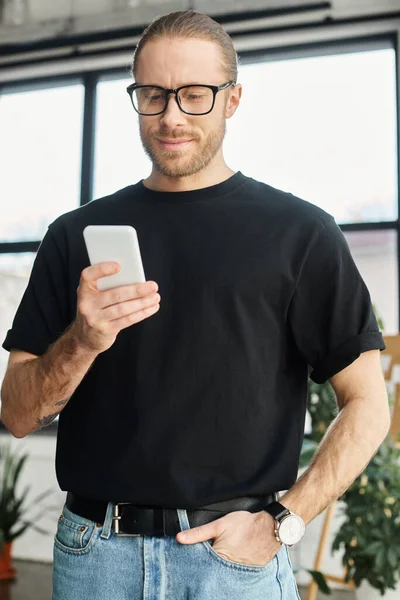 Uomo d'affari sorridente in occhiali e t-shirt nera con mano in tasca chatta su smartphone — Foto stock