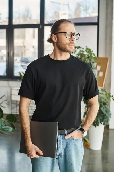 Gérant d'entreprise en t-shirt noir et lunettes debout avec ordinateur portable et la main dans la poche au bureau — Photo de stock
