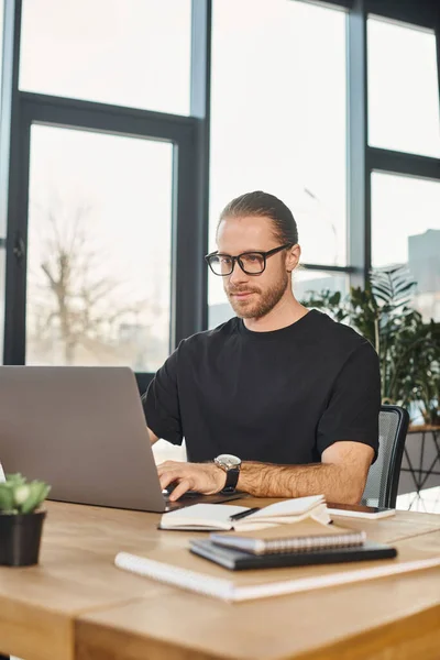 Стильный мужчина в черной футболке и очках, печатающий на ноутбуке на рабочем столе в современном офисе, бизнес — стоковое фото