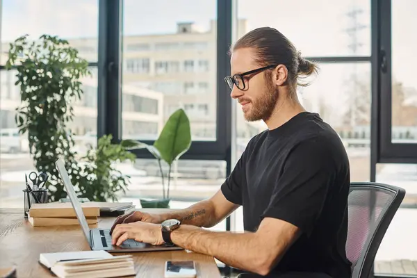 Hombre de negocios sonriente en camiseta negra y anteojos escribiendo en el ordenador portátil en el lugar de trabajo en la oficina moderna - foto de stock