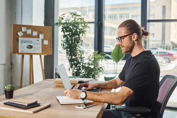 Side view of positive businessman in black t-shirt and eyeglasses working on laptop in office — Stock Photo