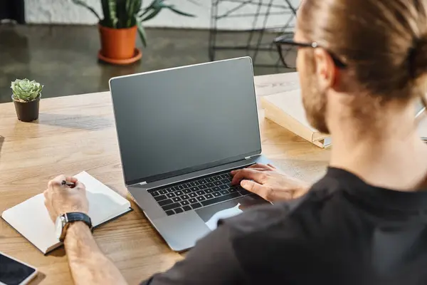 Бізнесмен в чорній футболці, що друкує на ноутбуці з порожнім екраном і пише в блокноті, вид ззаду — стокове фото