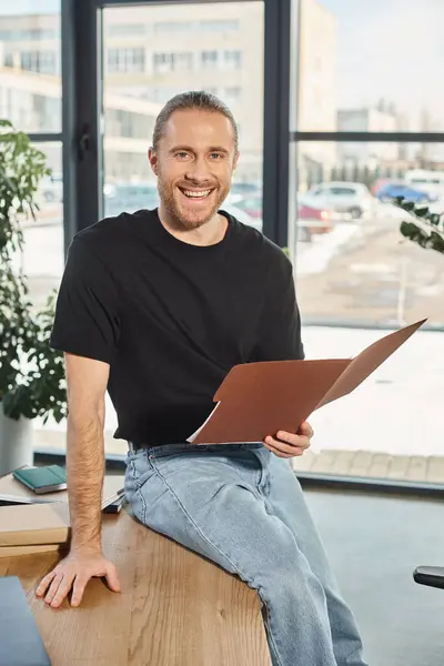Homme d'affaires souriant avec des lunettes assis sur le bureau avec un dossier et souriant à la caméra dans le bureau — Photo de stock