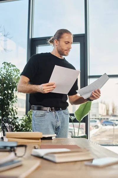 Uomo d'affari in abbigliamento casual guardando i documenti mentre in piedi e pensando alla scrivania in ufficio — Foto stock