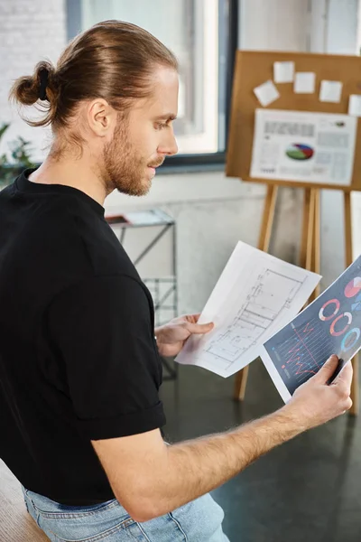 Homme d'affaires élégant en t-shirt noir regardant les papiers avec des graphiques et un plan dans le bureau moderne — Photo de stock