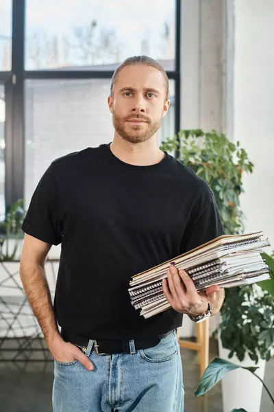 Task-oriented businessman in black t-shirt with hand in pocket holding pile of notebooks in office — Stock Photo