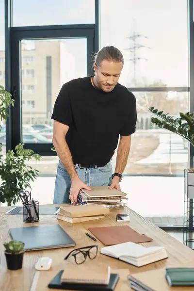 Trendy businessman in black shirt holding stack of notebooks while standing at work desk in office — Stock Photo