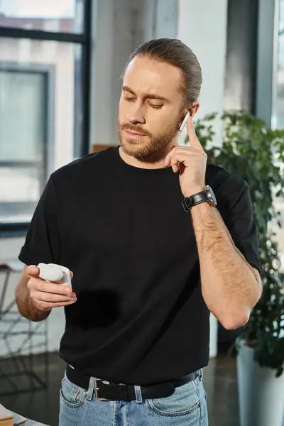 Hombre de negocios barbudo con estilo en camiseta negra insertando auriculares inalámbricos mientras está de pie en la oficina - foto de stock