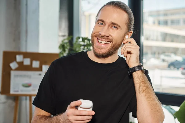 Cheerful businessman in casual attire adjusting wireless earphone and smiling at camera in office — Stock Photo