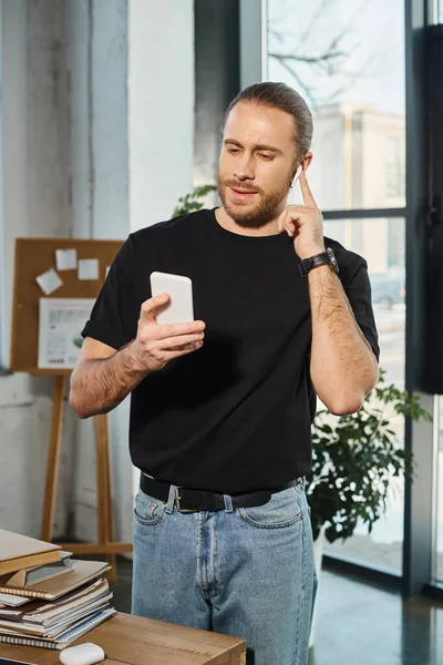Homme d'affaires élégant regardant smartphone et insérer un écouteur sans fil dans le bureau moderne — Photo de stock