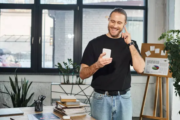 Homme d'affaires joyeux en t-shirt noir tenant smartphone et écouter de la musique dans les écouteurs au bureau — Photo de stock