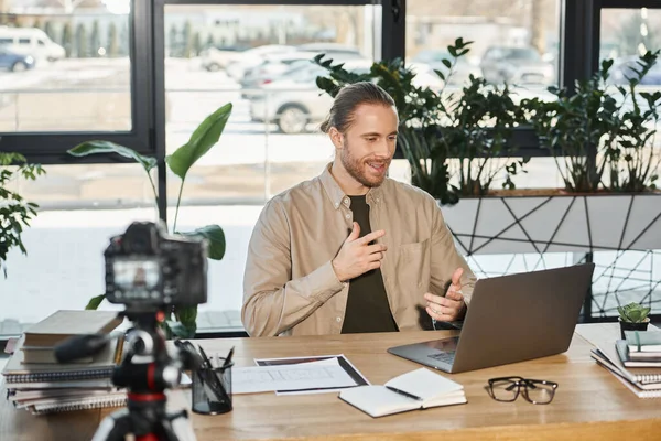 Erfolgreicher Geschäftsmann im Gespräch in der Nähe von Laptop und Digitalkamera während seines Videoblogs im Büro — Stockfoto