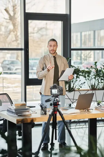 Uomo d'affari in abbigliamento casual in piedi con documento vicino alla fotocamera digitale e laptop, video marketing — Foto stock