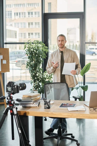 Seriöser Geschäftsmann mit Dokument und Videoblog vor Digitalkamera und Laptop — Stockfoto
