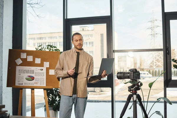 Бизнесмен с ноутбуком говорит рядом флип-чарт с аналитикой и цифровой камерой в современном офисе — стоковое фото