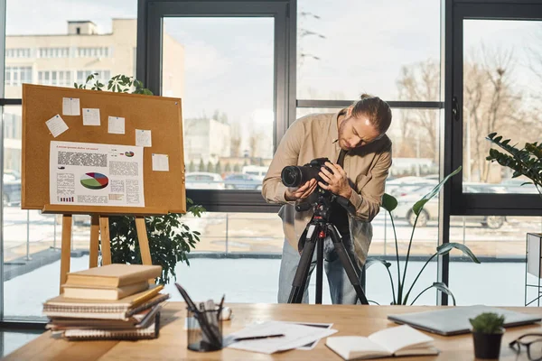 Kreativer Geschäftsmann justiert Digitalkamera in der Nähe von Flipchart mit Analytik und Schreibtisch im Büro — Stockfoto