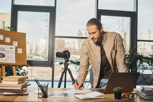 Geschäftsmann in Freizeitkleidung schreibt Notizen in der Nähe von Digitalkamera und Laptop auf Schreibtisch im Büro — Stockfoto