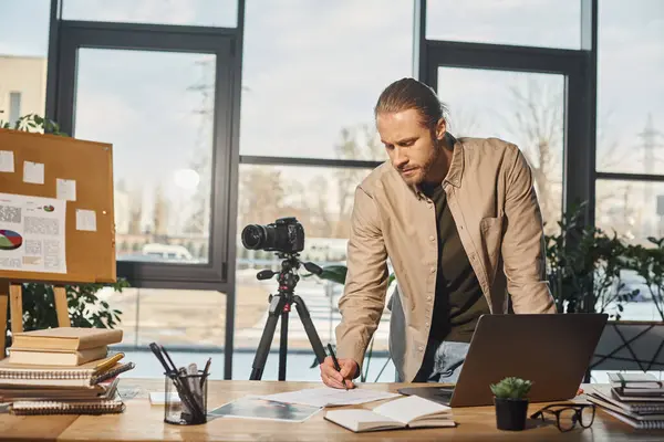 Businessman in casual attire writing notes near digital camera and laptop on work desk in office — Stock Photo