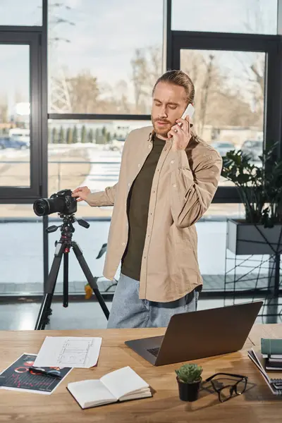 Homem de negócios falando no smartphone perto da câmera digital e laptop na mesa de trabalho no escritório moderno — Fotografia de Stock