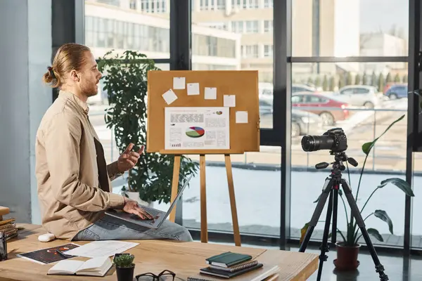 Homem de negócios com laptop sentado na mesa de trabalho perto da câmera digital e flip chart no escritório moderno — Fotografia de Stock