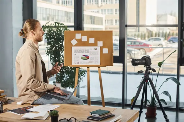 Homme d'affaires avec ordinateur portable assis sur le bureau près de l'appareil photo numérique et tableau à feuilles mobiles dans le bureau moderne — Photo de stock