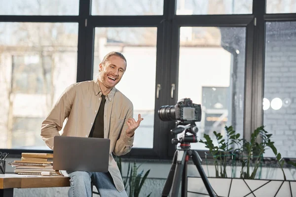Весёлый бизнесмен с ноутбуком разговаривает и жестикулирует возле цифровой камеры в современном офисе — стоковое фото